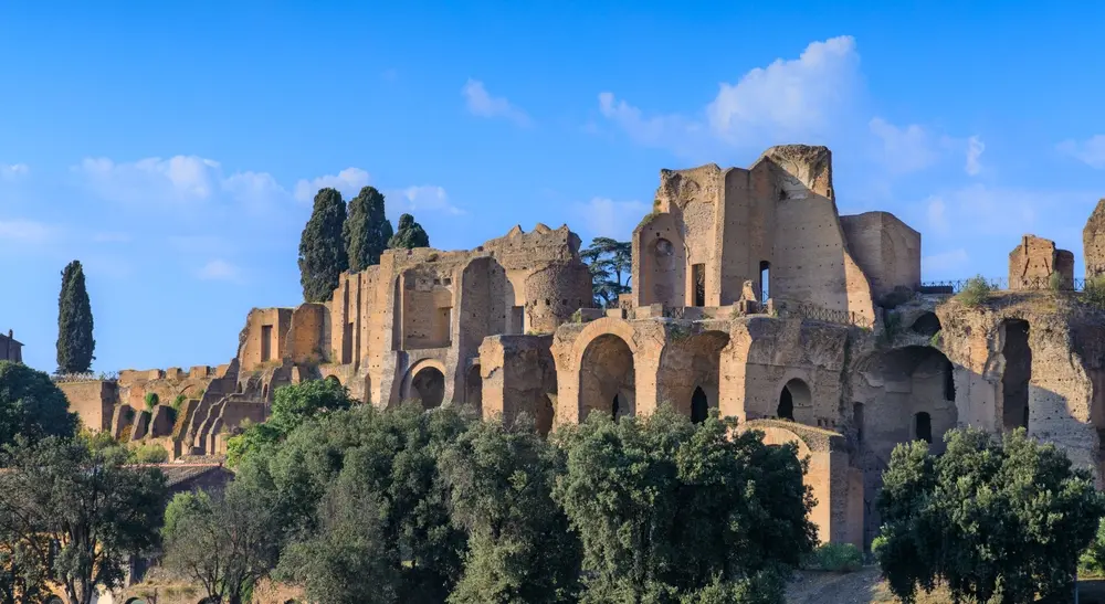 Ruins of the Palatine Hill in Rome, with ancient brick arches and walls rising above green trees against a blue sky